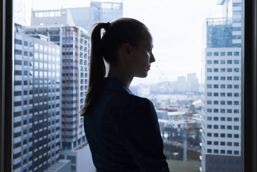 マンションの窓際に立ち、物憂げに階下を眺める外国人女性