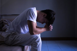 暗闇の中、ベッドに腰かけ落ち込むアジア人男性