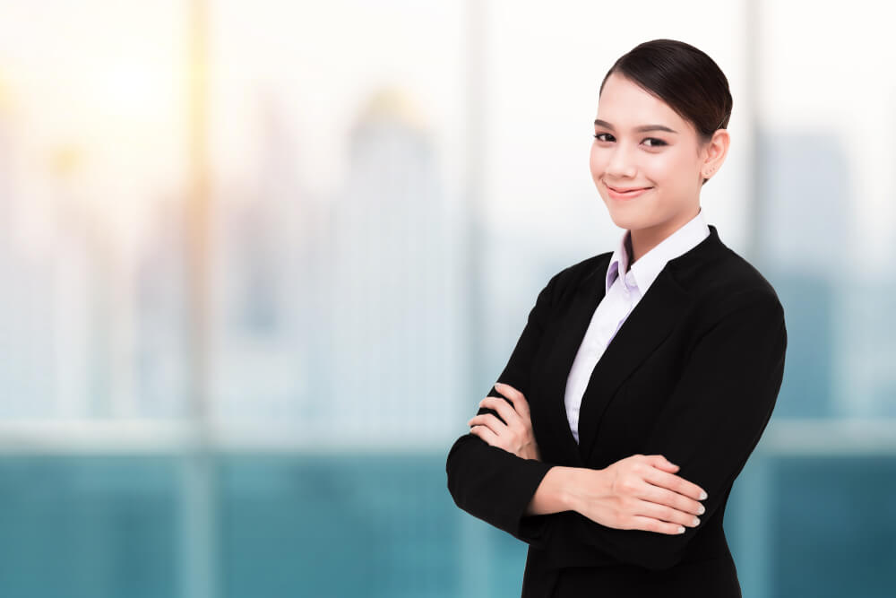 近代的オフィスの窓際で、腕を組んでこちらに微笑みかけるスーツ姿の外国人女性